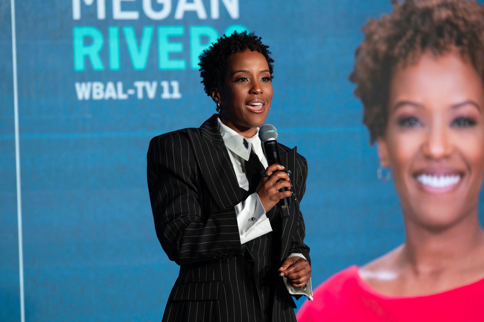 Megan Rivers speaking on stage at the MRCIAA event in a pinstripe suit