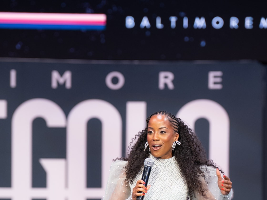 Megan Rivers speaking on stage in a white gown at the Baltimore Met Gala